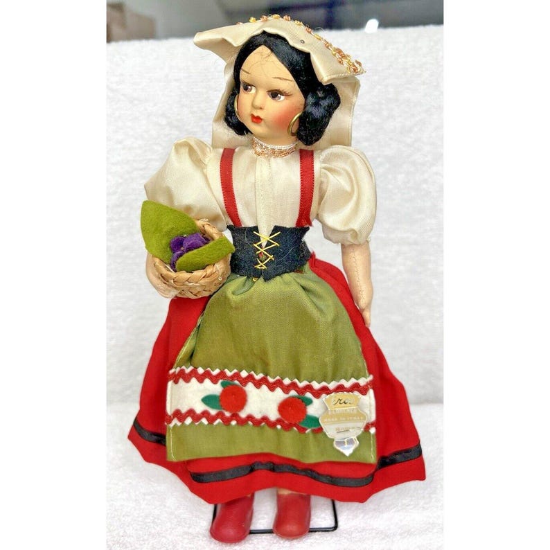 Peut inclure: Une poup&eacute;e vintage v&ecirc;tue d'une robe traditionnelle italienne avec un chemisier blanc, une jupe rouge et verte et un chapeau blanc. La poup&eacute;e tient un panier de fleurs violettes.