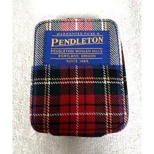 Puede incluir: Un artículo rectangular con un patrón de cuadros con una etiqueta azul que dice "PENDLETON WOOLEN MILLS, PORTLAND, OREGON, SINCE 1863". El diseño de cuadros presenta colores rojo, verde, negro y blanco. El artículo es probablemente un estuche pequeño o una cartera.