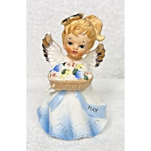 May include: A porcelain figurine of an angel with blonde hair, blue eyes, and a halo. She is wearing a white dress with blue trim and holding a basket of flowers. The dress has a blue ribbon with the word "MAY" on it.