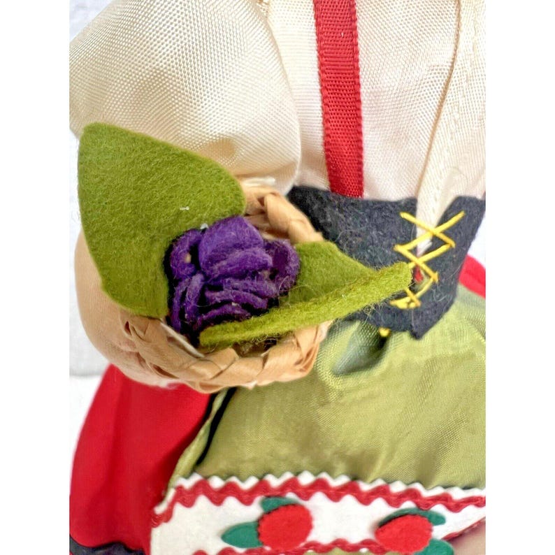Peut inclure: Une poup&eacute;e v&ecirc;tue d'un costume folklorique traditionnel avec une jupe rouge et verte, un chemisier blanc avec un ruban rouge et une ceinture noire avec un motif en croix. La poup&eacute;e tient un panier avec une fleur violette et des feuilles vertes.