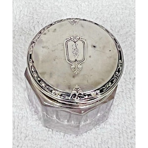 May include: A silver-toned, round-lidded jar with an ornate border and a decorative emblem on the lid. The jar's body is a clear, faceted glass. The lid features an engraved monogram. The jar is likely vintage or antique.