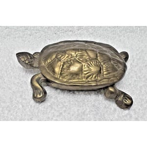 May include: A small, antique brass turtle figurine. The turtle has a detailed shell pattern and is resting on four small legs. The figurine is made of a shiny, golden-colored metal.