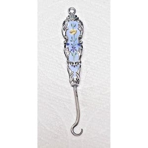Peut inclure: Un crochet en argent avec un manche décoratif. Le manche présente un motif émaillé bleu clair avec des accents floraux jaunes et violets. Le crochet a une extrémité incurvée.