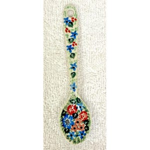 May include: A ceramic spoon featuring a cream-colored base with a hand-painted floral pattern. The handle is decorated with small blue flowers and green leaves, while the bowl displays a cluster of larger red, blue, and peach-colored flowers.