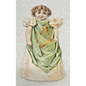 May include: A vintage paper doll featuring a young girl with curly brown hair and blue eyes. She wears a light green dress with a gold harp design. The doll has puffed sleeves and a long skirt, with a cream-colored background.