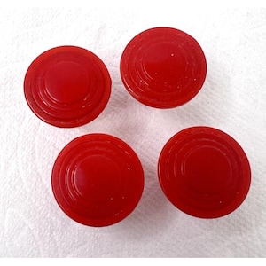 May include: Four vibrant red, circular game pieces with concentric ring detailing. The pieces are evenly spaced against a white background. These could be checkers, game tokens, or other board game components.