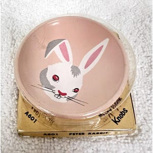 May include: A round, light pink knob featuring a white rabbit illustration with gray ears, red eyes, and black whiskers. The packaging is a gold-colored square base with clear plastic, labeled with the text A601 and Peter Rabbit Knobs.
