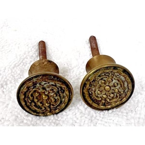 May include: Two antique brass cabinet knobs with ornate, oval faces. The knobs have a detailed floral and scroll design, with a slightly aged, tarnished appearance. Each knob has a threaded post for mounting.