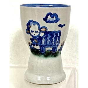 May include: A ceramic egg cup with a speckled off-white glaze and a blue rim. The exterior features a hand-painted blue illustration of a sheep standing on a patch of green grass, with a blue cloud shape above the sheep's head.
