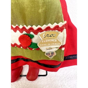 Peut inclure: Une poup&eacute;e rouge et verte portant une jupe rouge avec une bordure noire, une jupe verte avec une bordure blanche et rouge, et un pompon blanc avec un centre rouge. La poup&eacute;e porte une &eacute;tiquette qui indique "Eros Florence Made in Italy".