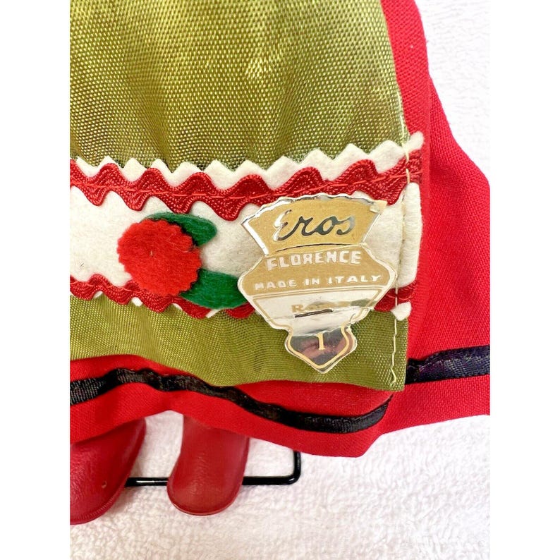 Peut inclure: Une poup&eacute;e rouge et verte portant une jupe rouge avec une bordure noire, une jupe verte avec une bordure blanche et rouge, et un pompon blanc avec un centre rouge. La poup&eacute;e porte une &eacute;tiquette qui indique "Eros Florence Made in Italy".