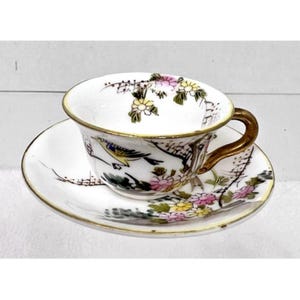 May include: A white porcelain teacup and saucer set with a floral and bird design. The cup and saucer have a gold trim and are decorated with pink, yellow, and green flowers and a yellow bird.