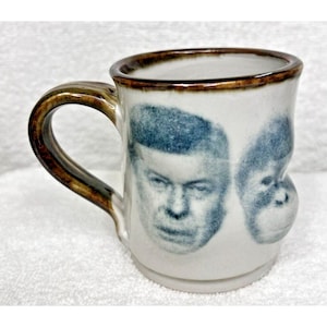 May include: A white ceramic mug with a brown handle. The mug has a black and white image of a man's face and a monkey's face on the front.