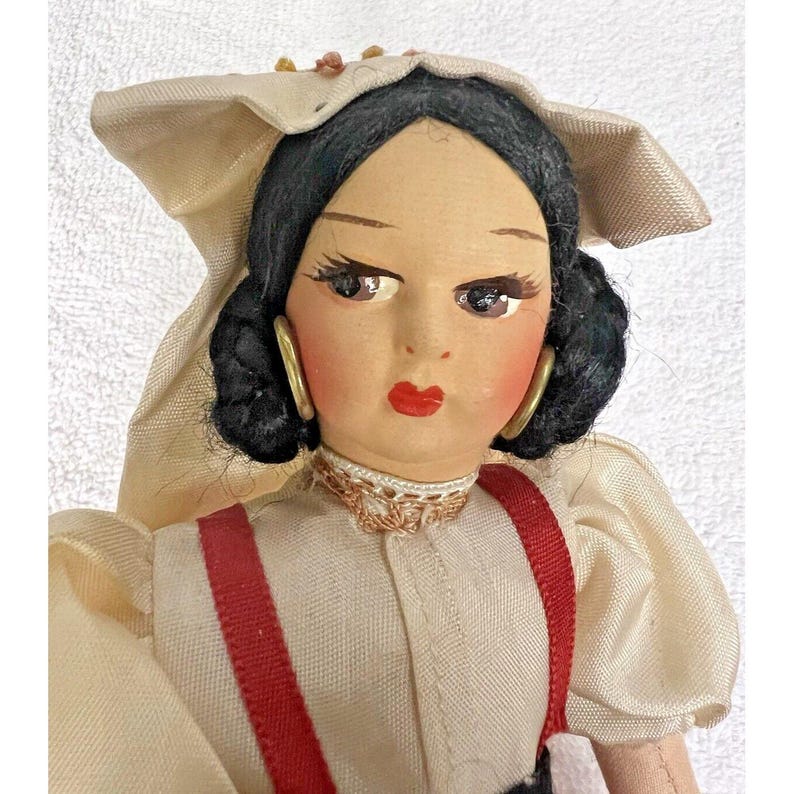 Peut inclure: Une poup&eacute;e vintage aux cheveux noirs, portant une robe blanche avec des bretelles rouges et une coiffe blanche. La poup&eacute;e a des traits peints et porte des boucles d'oreilles dor&eacute;es.