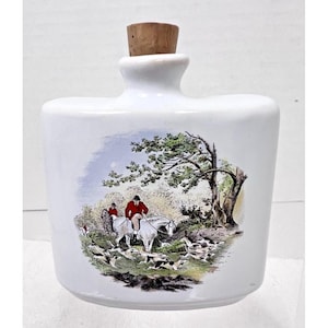 May include: White ceramic bottle with a cork stopper and a hunting scene illustration. The scene depicts two people on horseback with hounds running through a forest.