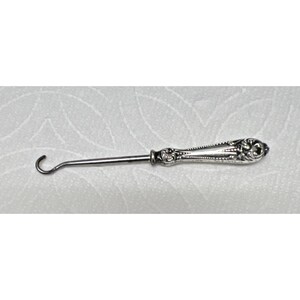 May include: A silver-toned antique crochet hook with a curved end and ornate handle. The handle features floral and scroll designs, with a textured, detailed surface. The hook is designed for crafting and textile work.