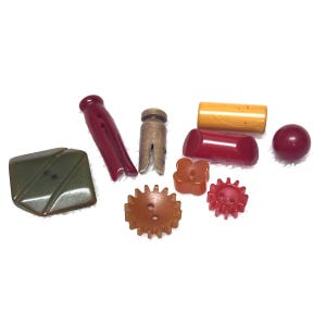 May include: A collection of assorted sewing buttons and fasteners including a large olive green diamond-shaped button, a maroon clothespin-style fastener, a metallic cylinder, a yellow rectangular bar, a small red sphere, and gear-shaped buttons.