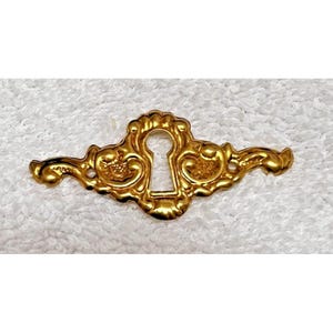 May include: A gold-colored keyhole cover with ornate scrollwork and a keyhole-shaped opening. The decorative hardware has a symmetrical design with curved edges and a polished finish. This vintage-style piece is likely for furniture.