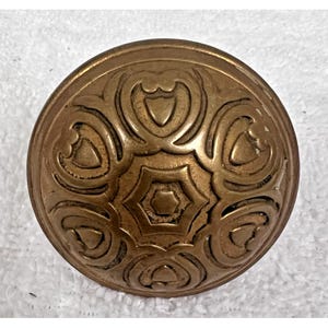 May include: A round, antique brass button with an intricate, embossed floral design. The button features a central six-pointed star surrounded by stylized heart and swirl patterns. The button is slightly domed and has a warm, aged patina.