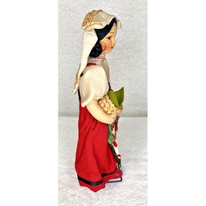 Peut inclure: Une poup&eacute;e vintage portant un foulard blanc avec des accents dor&eacute;s, un chemisier blanc et une jupe rouge. La poup&eacute;e tient un panier avec des feuilles vertes.