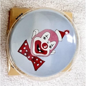 May include: A round, clear plastic button featuring a light blue background with a graphic illustration of a clown face. The clown has a pink face, a red nose, a red mouth with visible teeth, and a red bow tie with white polka dots.