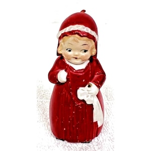 May include: A vintage ceramic figurine of a young girl in a red dress and matching hat with white trim. The figurine is holding a white cloth and has a painted face with blonde hair.