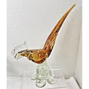 Puede incluir: Escultura de vidrio de un gallo con vidrio ámbar y transparente. El gallo tiene una cola larga y curvada y una cresta detallada. La escultura está montada sobre una base de vidrio transparente.