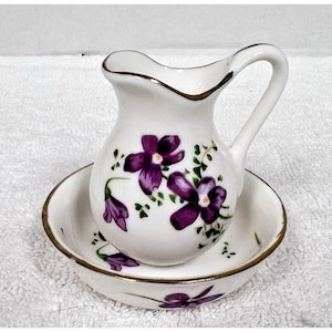 May include: A white porcelain miniature pitcher and bowl set. The set is decorated with purple floral designs and gold trim. The pitcher has a handle and a spout. The bowl is round and sits beneath the pitcher.
