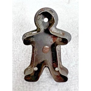 May include: Vintage metal gingerbread man cookie cutter. The cutter is made of a dark, aged metal with visible rust spots. The design features a classic gingerbread man shape with arms, legs, and a head with a hole at the top. A small, round indentation is in the center.