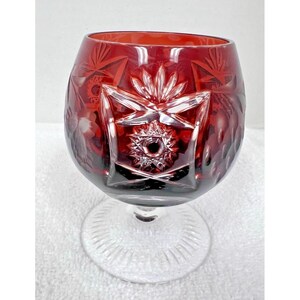 May include: A ruby red crystal glass with intricate starburst and floral cut designs. The glass has a rounded bowl and a clear, etched stem and base. This decorative glassware is ideal for serving beverages.