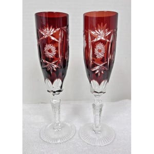 May include: Two ruby red crystal champagne flutes. Each glass features a tall, slender bowl with intricate starburst and floral cut designs. The stems are clear and detailed, resting on a circular base. The glasses are displayed against a white background.