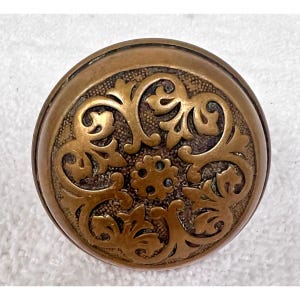 May include: A round, antique bronze button with an ornate floral design. The button features a central flower surrounded by swirling leaves and vines. The button has a slightly domed shape and a detailed, textured surface.