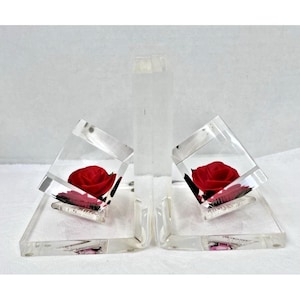 May include: Clear acrylic bookends with a red rose encased in each angled cube. The bookends have a rectangular center piece and a triangular base. The roses are a vibrant red color, and the acrylic is transparent.