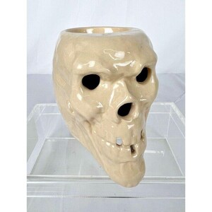 May include: A beige ceramic skull-shaped container with black eye and nose holes. The skull has a detailed, textured surface and a wide opening at the top. The container is likely used for decorative purposes.
