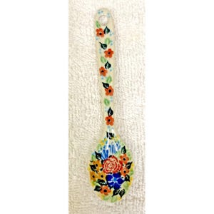 May include: A ceramic spoon featuring a cream-colored base with a hand-painted floral pattern. The handle displays small orange flowers and green leaves, while the bowl of the spoon showcases a larger cluster of red, blue, and orange flowers.