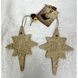 May include: Two beige ceramic star ornaments featuring an embossed floral pattern. Each star hangs from a string adorned with wooden beads. A rectangular tag attached to the string displays the text Matthew Kelly Pottery and an image of a vase.