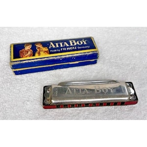 Può includere: Un'armonica vintage e la sua scatola. L'armonica è argentata con la scritta "ATTA BOY" e dettagli rossi. La scatola è blu con la scritta "ATTA BOY" e un'illustrazione di due persone che suonano l'armonica. Prodotto da F.R. HOTZ Germania.