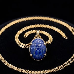May include: A gold chain necklace featuring an oval-shaped lapis lazuli pendant carved into a scarab beetle design. The deep blue stone displays natural gold-colored flecks and is secured in a gold setting with four prongs at the edges.
