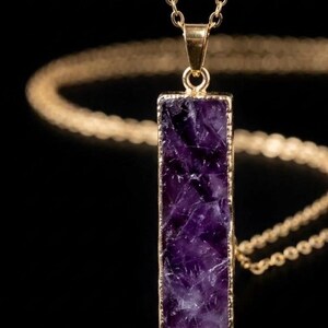 Peut inclure: Un pendentif rectangulaire en améthyste violette à la surface texturée et irrégulière, serti dans un fin cadre en métal doré. Le pendentif est suspendu à une délicate chaîne dorée, dotée d'une bélière polie reliant la pierre au collier.