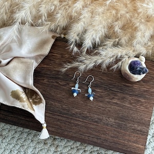 May include: Dangle earrings with silver hooks, each featuring blue and clear beads, and a small white bead. The earrings are presented on a dark wooden surface. The earrings are a delicate piece of jewellery.