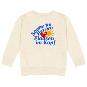 Puede incluir: Sudadera de color crema con mangas largas. La parte trasera de la sudadera presenta el texto "Sonne im Herzen, Flausen im Kopf" en azul, con un corazón rojo y un sol amarillo. La sudadera está hecha de un material suave y cómodo.