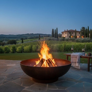 Può includere: Un grande braciere circolare in metallo marrone scuro con una forma a coppa ampia e poco profonda. Il braciere è riempito con ceppi ardenti che producono fiamme arancioni e gialle brillanti, posizionato su una superficie di patio in pietra.