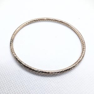 May include: A silver-colored bangle bracelet with a textured, etched surface. The bracelet is a simple, circular design, suitable for everyday wear. The bangle is shown against a white background.