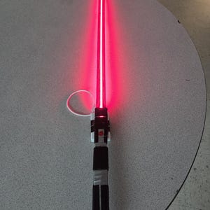May include: A red lightsaber with a glowing blade. The hilt is black, white, and silver with a rectangular design. The lightsaber is on a gray surface.