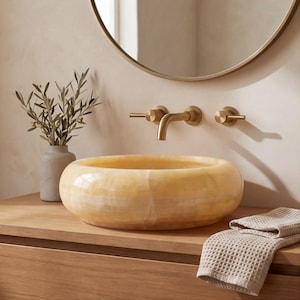 May include: A round, polished yellow onyx stone vessel sink with natural banding, paired with a wall-mounted brushed brass faucet featuring two lever handles, set on a wooden vanity with a beige waffle-weave towel and a small ceramic vase.