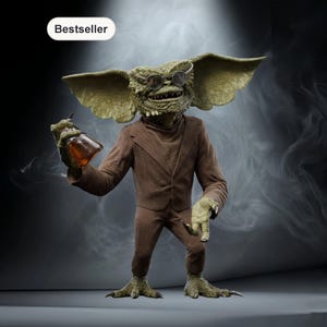 Puede incluir: Figura detallada de un Gremlin con traje marrón y gafas redondas pequeñas, sosteniendo una botella marrón. La figura tiene orejas grandes y una cara verde texturizada. La palabra "Bestseller" está en un óvalo blanco en la esquina superior izquierda.