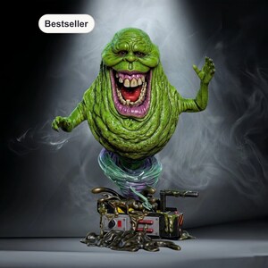 May include: A green, smiling, cartoon-like figure with a large mouth and teeth, standing on a base with a device and slime. The figure has its arms raised. The word "Bestseller" is in a white oval in the upper left corner.