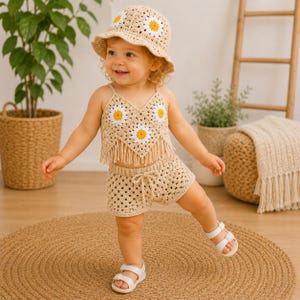 May include: A beige crochet outfit consisting of a bucket hat, a V-neck crop top with fringe, and shorts. The hat and top feature white and yellow daisy motifs. The shorts have a drawstring waist. The set is made of a light tan yarn material.