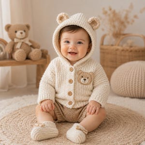 May include: A smiling baby wearing a cream-colored knitted bear hoodie with matching booties and shorts. The hoodie has bear ears and a bear face patch. The baby is sitting on a round, woven rug.