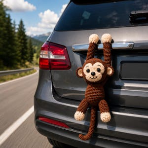 Könnte beinhalten: Ein gehäkelter Affe in Braun- und Beigetönen, der an der Rückseite eines grauen Autos hängt. Der Affe hat ein lächelndes Gesicht und schwarze Augen. Das Auto steht auf einer Straße mit Bäumen und Bergen im Hintergrund.